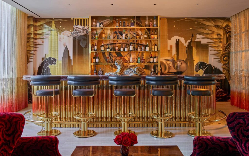 Faena Hotel bar with a gold ribbed counter, black stools, an Art Deco-style mural, and a spotted cat sculpture among the liquor shelves.