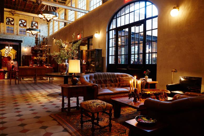 Hotel Emma lobby with leather seating, patterned tile floors, large arched windows, candlelit tables, and preserved brewery machinery in the background.