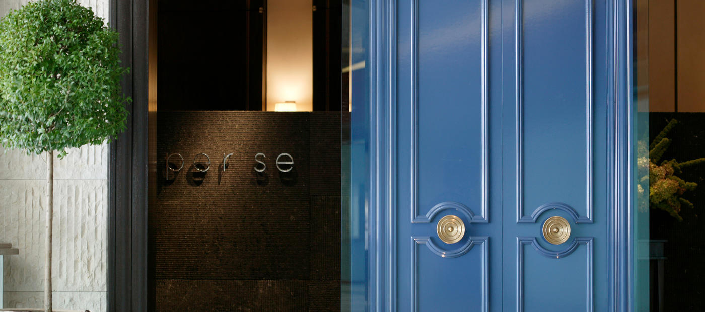 Per Se entrance with deep blue double doors, brass round handles, a clipped topiary, and the restaurant name on a dark wall.