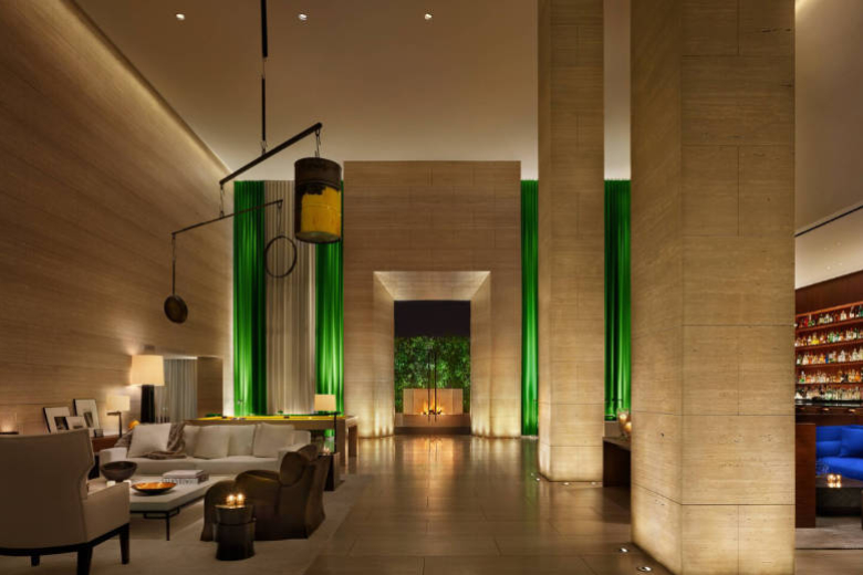 West Hollywood EDITION lobby with monumental stone columns, tall green drapery, a central fireplace, neutral lounge seating, and a bar at the right.