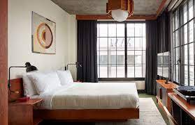 Ace Hotel guest room with a wood platform bed, white linens, concrete ceiling, and large black-framed industrial windows.