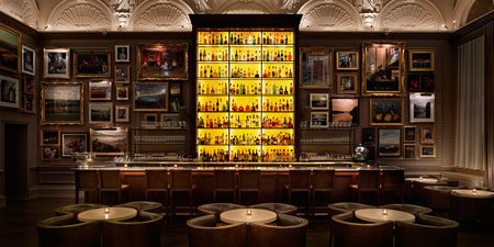 London EDITION bar with a tall illuminated wall of bottles, dark wood counter, salon-style framed artwork, and intimate round seating.