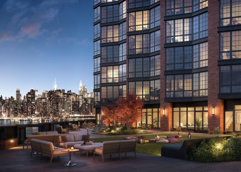 Greenpoint Landing terrace with lounge seating and landscaped lawn beside a brick-and-glass tower, overlooking the Manhattan skyline at dusk.