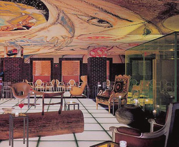 Hudson Hotel lounge with an expansive abstract painted ceiling, mixed vintage seating, brick columns, and geometric white floor tiles.