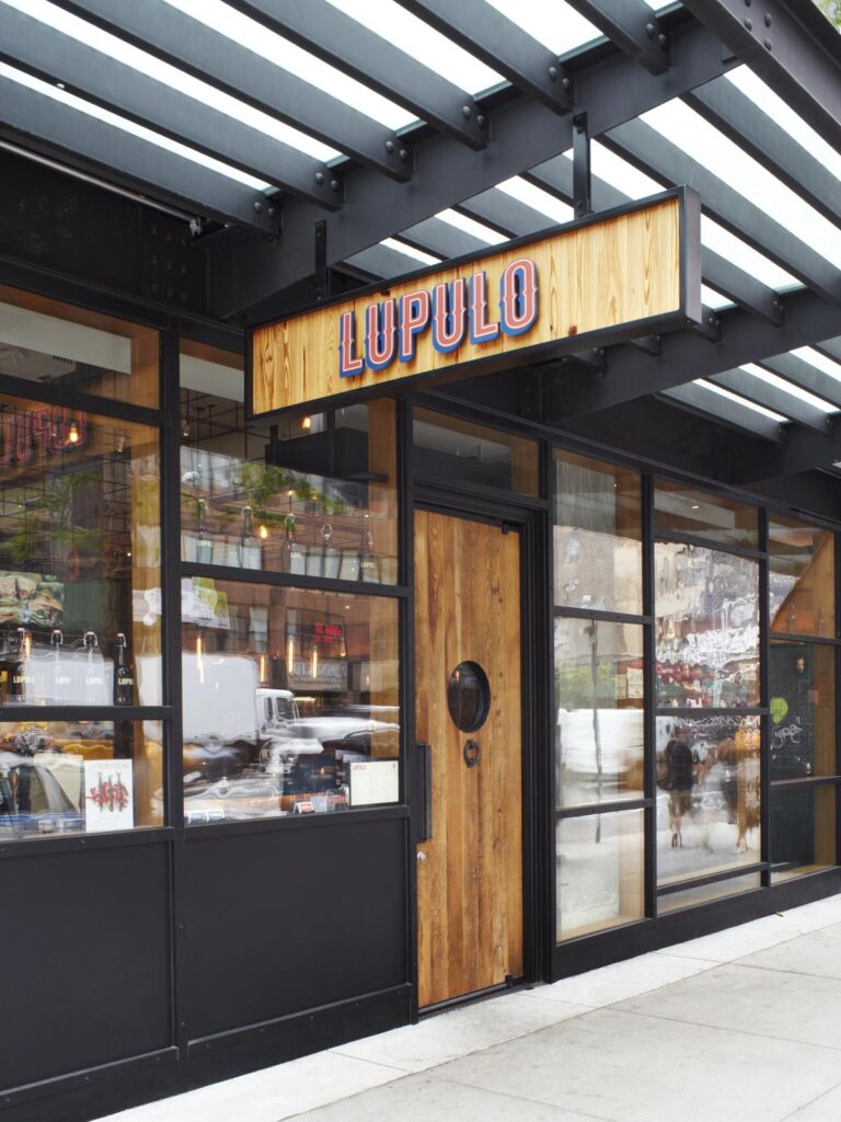 Lupulo restaurant storefront with black-framed glass windows, a wood entry door, and an illuminated LUPULO sign beneath a metal canopy.