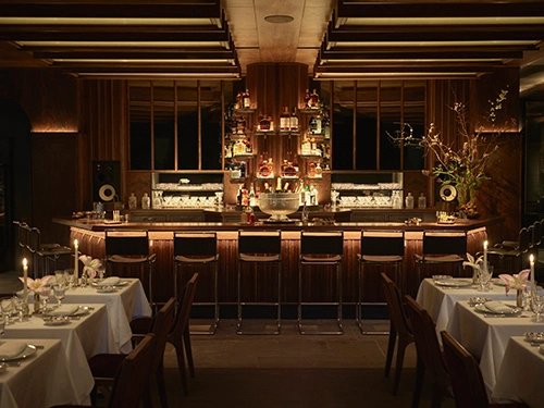 Warmly lit Marcel restaurant interior with a dark wood bar, six stools, mirrored liquor shelves, and white-tablecloth dining tables.