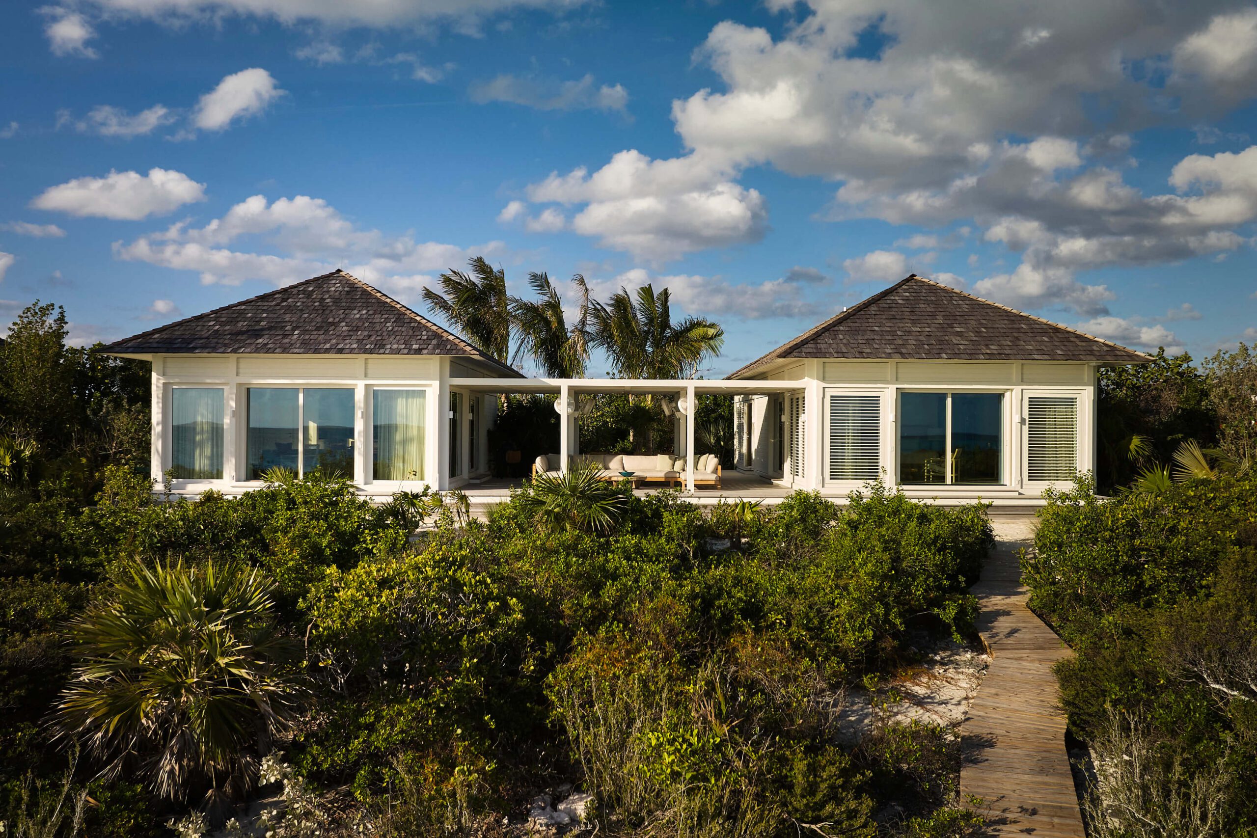 Norman’s Cay villa with two white pavilion wings, dark shingled roofs, palm trees, and a wooden path through tropical landscaping.