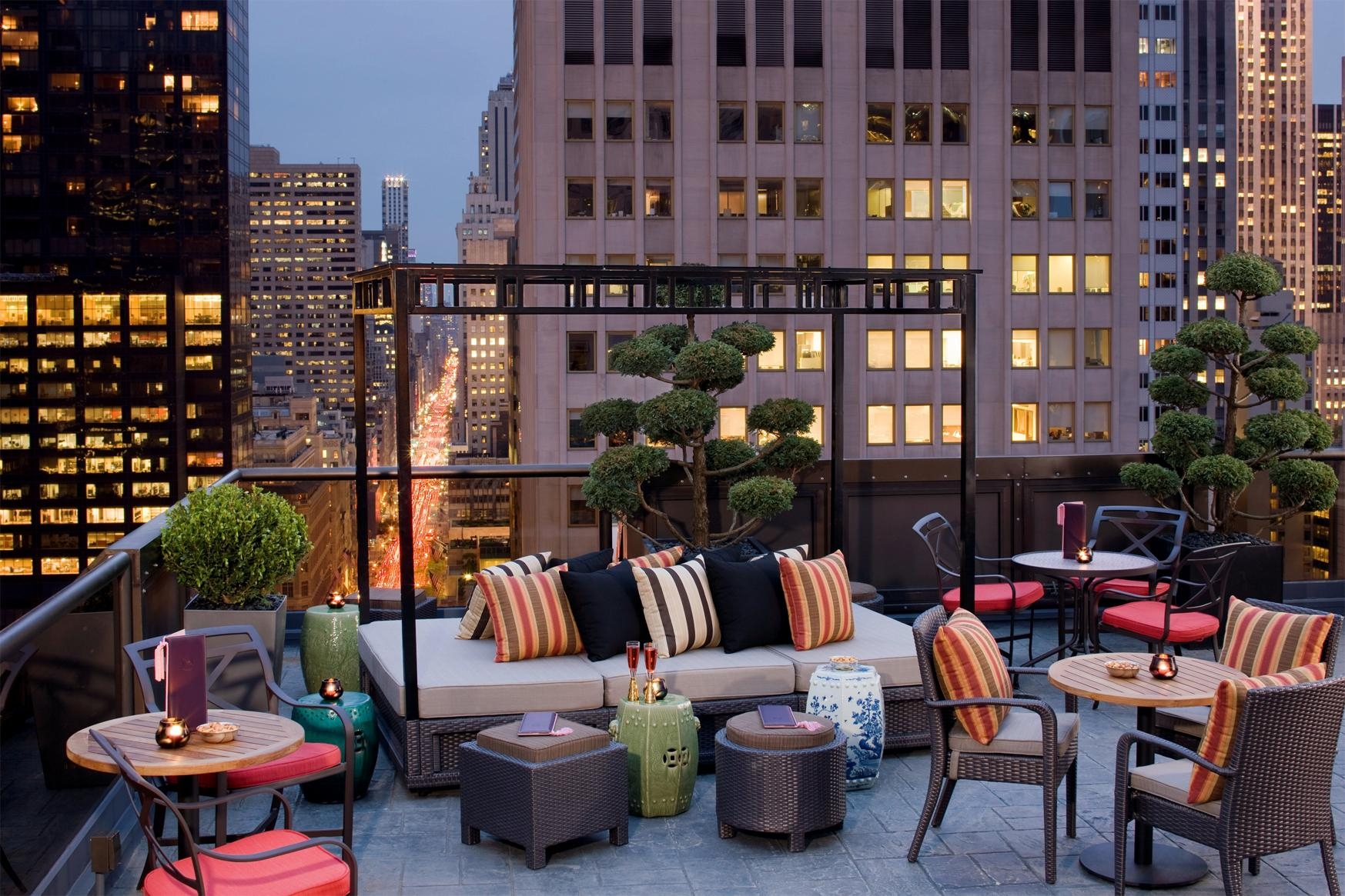 Peninsula rooftop terrace at dusk with wicker seating, striped cushions, sculpted topiary, and illuminated Manhattan buildings.