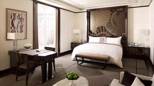 Peninsula Paris guest room with a white bed, dark decorative headboard, writing desk, tall window, and understated neutral furnishings.