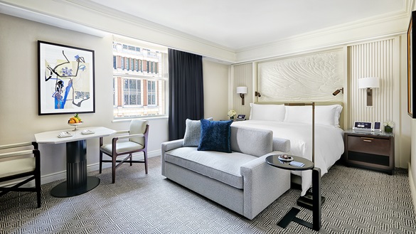 Peninsula New York guest room with a white bed, gray sofa, blue accent pillow, dining table, and framed artwork beside a large window.