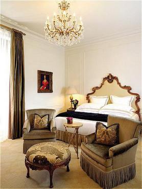 Plaza Hotel guest room with an ornate headboard, crystal chandelier, upholstered armchairs, and traditional cream-and-gold decor.