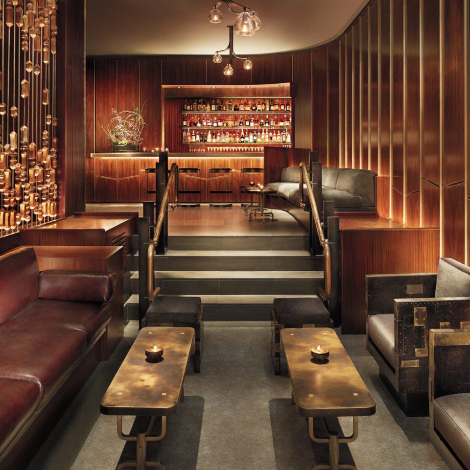 Royalton Hotel lounge with rich wood walls, leather seating, bronze tables, a raised bar, and sculptural hanging lights.
