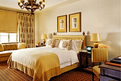 St. Regis Washington guest room with cream bedding, gold-striped upholstery, patterned drapes, framed artwork, and a decorative chandelier.
