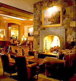 St. Regis Aspen lounge with a large stone fireplace, wood-paneled walls, patterned armchairs, and a framed painting above the hearth.