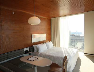 Standard Hotel guest room with wood-paneled walls and ceiling, a white bed, built-in banquette, oval table, and large city-facing window.