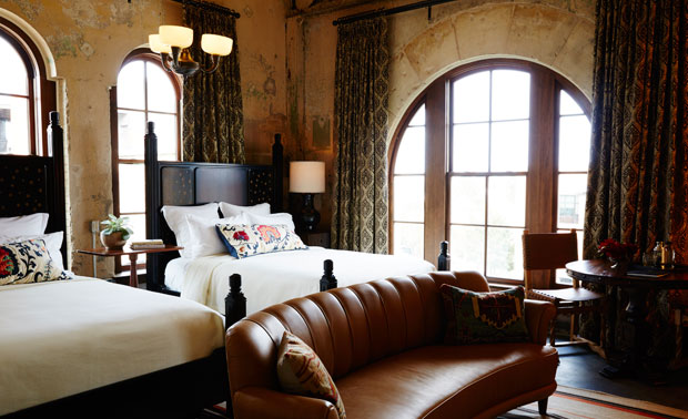 Hotel Emma guest room with two beds, tall arched windows, weathered plaster walls, patterned curtains, and a brown leather sofa.