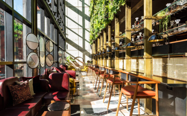 Moxy Hotel conservatory bar with tall windows, burgundy lounge seating, a long brass-accented bar, and hanging greenery.