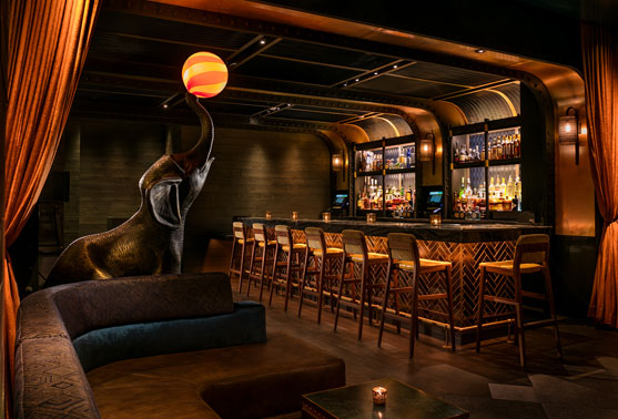 Moxy Hotel bar with a large elephant sculpture balancing a glowing orb beside a dark wood bar and rows of stools.