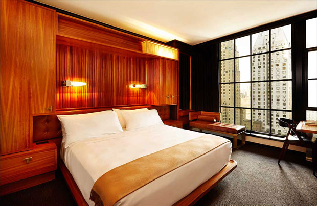 Viceroy Hotel guest room with warm wood paneling, a white bed, and a floor-to-ceiling grid window overlooking neighboring skyscrapers.