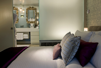 W Hotel guest room with white bedding and purple accent pillows, with a bright mirrored bathroom visible beyond the bed.